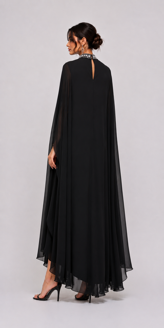 A woman modeling a long black evening gown with a sheer cape-like overlay and a small keyhole cutout at the back, shown from behind against a light gray studio background.