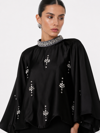A woman wearing a black satin cape-style top with silver crystal embellishments on the collar and scattered across the front, photographed against a light gray studio background.