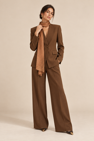 A model wearing a brown tailored pantsuit with a matching scarf, styled with pointed shoes, against a solid beige studio background.