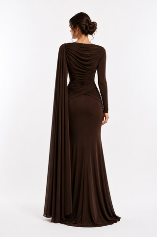 A woman viewed from the back wearing a floor-length dark brown gown with long sleeves, draped neckline, and a flowing cape-like side drape, photographed against a clean light gray studio background.