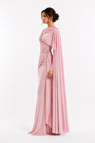 A woman modeling a floor-length dusty pink evening gown with draped ruching, long sleeves, and a flowing cape-like overlay, shown in side profile against a clean white studio background.