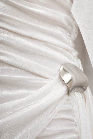 A close-up of a white draped fabric garment with gathered ruching and a large, polished silver metallic ring detail at the waist.