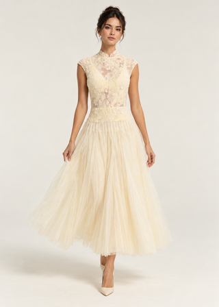 A woman models a sleeveless cream tulle dress with a lace bodice and high collar, standing against a plain light background.