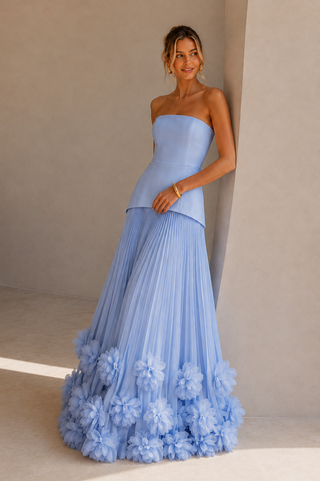A woman models a strapless light blue gown with a structured bodice and a voluminous pleated skirt adorned with large floral ruffles at the hem, posed against a minimalist beige wall.