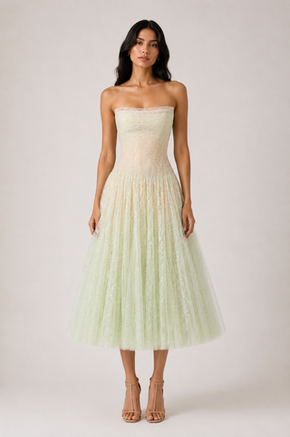 A woman modeling a strapless pale green tulle midi dress with a lace corset-style bodice, photographed against a plain light gray studio background.
