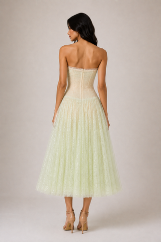 A woman shown from the back wearing a strapless pale yellow tulle dress with a structured corset-style bodice and a full pleated skirt, photographed against a plain warm beige studio background.