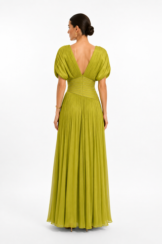 A woman viewed from the back wearing a floor-length olive green gown with puff sleeves, a deep V back, and a pleated skirt, photographed against a clean white studio background.