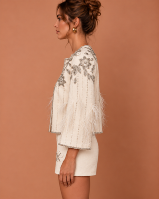 A woman shown in side profile wearing a white embellished jacket with silver beadwork, feathered trim, and fringed cuffs, paired with matching white shorts, against a solid terracotta background.