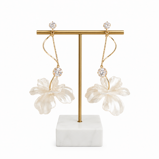 A pair of gold-tone dangle earrings with large white flower charms and clear crystal studs, displayed on a gold T-stand with a white marble base against a white background.