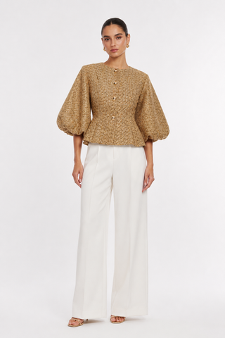 A model stands against a light gray studio background wearing a cropped, textured tan top with puff sleeves and white wide-leg trousers.