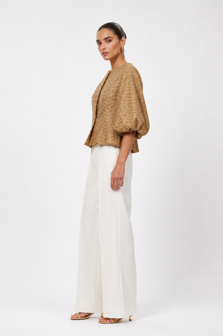A woman modeling a cropped, textured tan jacket with voluminous puff sleeves and white wide-leg trousers, standing against a plain white studio background.