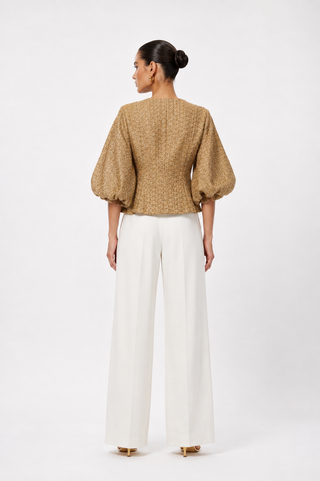 A woman is shown from the back wearing a textured tan top with voluminous puff sleeves and white wide-leg trousers, standing against a plain white studio background.