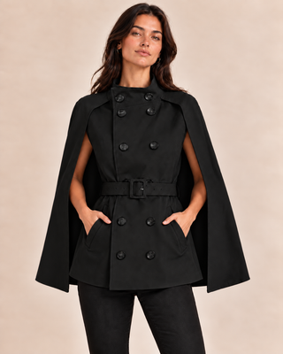 A woman modeling a black double-breasted cape jacket with a matching belt, shown against a plain beige studio background.