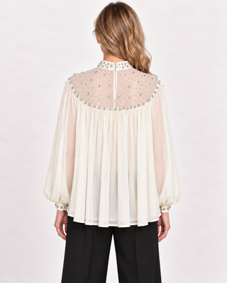 A woman viewed from the back wearing a cream-colored long-sleeve blouse with a sheer, embellished yoke and black trousers, set against a plain light background.
