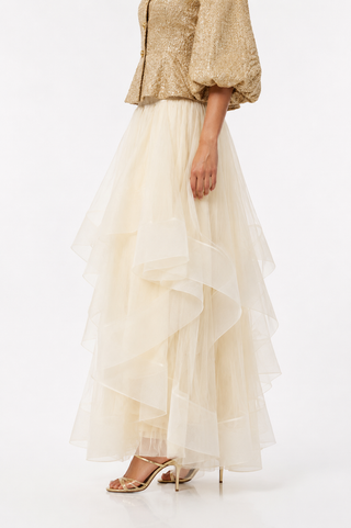 A model is shown from the waist down wearing a long, layered tulle skirt in cream/off-white, paired with a textured gold top and gold heeled sandals, against a plain white studio background.
