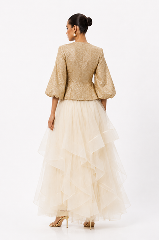A woman shown from the back wearing a formal evening gown with a textured champagne-gold cropped jacket and a layered cream tulle skirt, photographed against a plain white studio background.