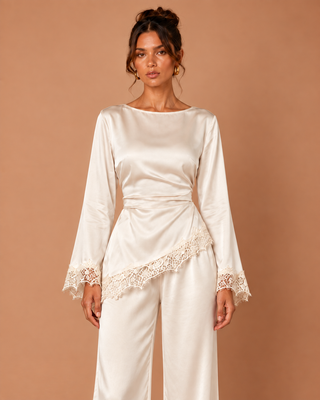 A woman modeling a cream satin long-sleeve top and matching wide-leg pants against a solid warm beige background. The top features a draped waist with a lace overlay and lace-trimmed cuffs.