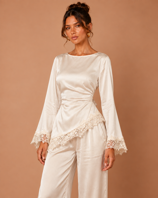 A woman modeling a cream satin long-sleeve top with lace trim and matching wide-leg pants against a solid warm beige studio background.