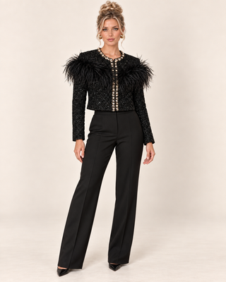 A woman models a black embellished jacket with dramatic feathered shoulders and black wide-leg trousers against a plain beige studio background.