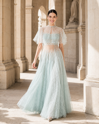 A woman wearing a light mint green lace and tulle two-piece dress stands in a sunlit stone colonnade. The dress has a sheer high-neck crop top with flutter sleeves and a matching full skirt.