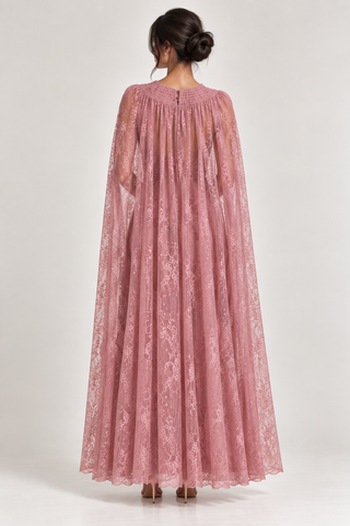 A woman viewed from the back wearing a long dusty pink lace cape dress with sheer sleeves, standing against a light gray studio background.