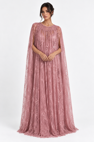 A woman modeling a floor-length dusty rose pink gown with a sheer cape-like overlay and intricate floral lace pattern, set against a plain white studio background.