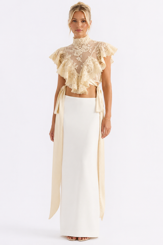 A model wearing a two-piece formal outfit featuring a cream lace ruffled crop top with side ties and a matching cream satin high-waisted skirt, photographed against a solid white studio background.