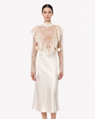 A model wearing a cream-colored dress featuring a sheer lace long-sleeve top with ruffles over a satin midi skirt, shot against a plain light gray background.
