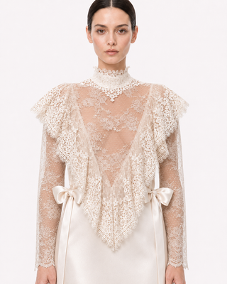 A model wearing a cream lace high-neck shrug with ruffled shoulders and bow-tied sleeves over a cream satin dress, set against a plain white studio background.