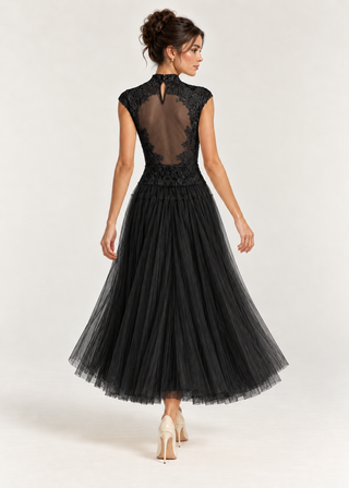 A woman modeling a black sleeveless midi dress from the back, featuring a sheer lace back panel, high neck, and a voluminous black tulle skirt, set against a plain light gray studio background.