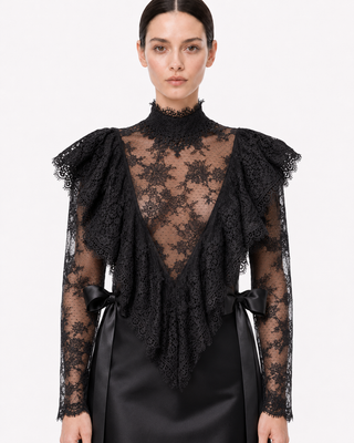 A model wearing a black lace mock-neck top with dramatic ruffled shoulders, sheer long sleeves, and a deep V-neckline, paired with a black satin skirt against a plain white studio background.