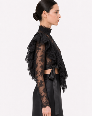 A model shown in profile wearing a black lace long-sleeve crop top with ruffled layers and a black satin skirt against a plain white studio background.