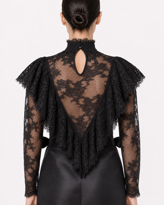 Back view of a model wearing a black lace long-sleeve top with a high mock neck, keyhole back cutout, and dramatic ruffled shoulders, paired with black satin trousers against a light gray studio background.