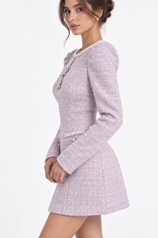 A woman wearing a light lavender tweed mini dress with pearl trim and a decorative pearl-and-gold collar detail, photographed against a clean white studio background.