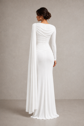 A woman modeling a floor-length white gown from the back, featuring a draped neckline with a long flowing capelet, fitted long sleeves, ruched detailing at the waist, and a slight train, set against a neutral textured studio backdrop.