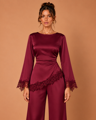 A woman modeling a matching long-sleeve top and wide-leg pants set in deep burgundy satin, featuring lace trim at the cuffs and a draped lace panel at the waist, against a solid warm tan studio background.