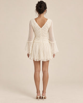 A woman viewed from the back wearing a short cream chiffon dress with long sheer sleeves, a ruched V-neck bodice, and a ruffled mini skirt, paired with beige high-heeled sandals against a plain beige studio background.