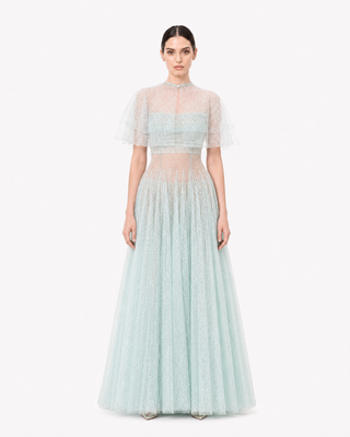 A model wearing a light mint green evening gown with a sheer mesh crop top, short flutter sleeves, and a full tulle skirt, photographed against a plain white background.