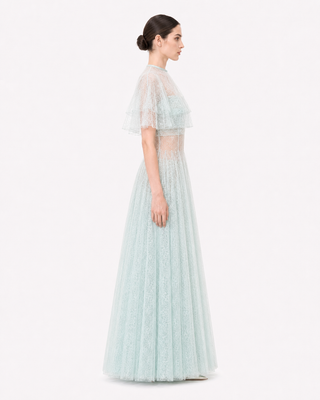 A woman modeling a floor-length light aqua lace gown with sheer flutter sleeves and a fitted waist, shown in side profile against a plain white studio background.