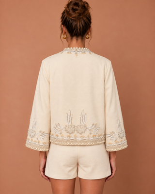 A woman shown from the back wearing a cream-colored embroidered jacket with matching shorts, set against a solid warm terracotta background.