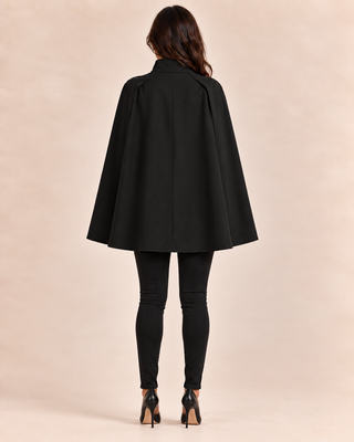 A woman viewed from the back wearing a black cape-style coat over black leggings and black high heels, standing against a plain warm beige studio background.