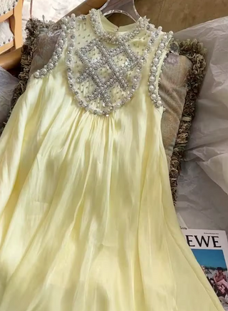A pale yellow sleeveless dress with a high round neckline, decorated with a large silver beaded geometric pattern across the chest and shoulders, shown hanging on a hanger.
