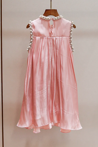 A sleeveless light pink satin dress hanging on a wooden hanger against a plain beige wall. The dress features a gathered waist and is decorated with white pearl-like embellishments along the shoulder straps and neckline.