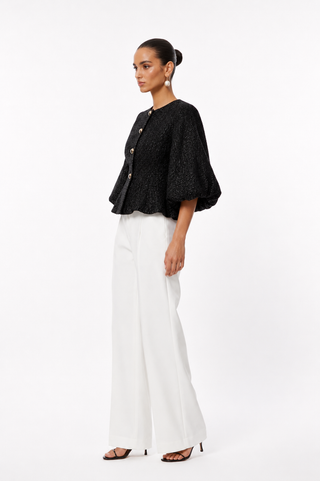 A woman modeling a black textured short-sleeve cropped jacket with gold buttons and flared sleeves, paired with white wide-leg trousers and black sandals, against a plain white studio background.