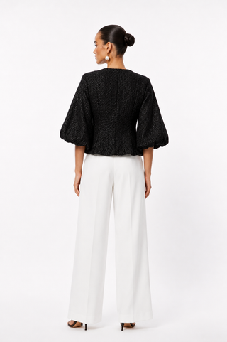 A woman shown from the back wearing a black textured puff-sleeve top and white wide-leg trousers, standing against a plain white studio background.