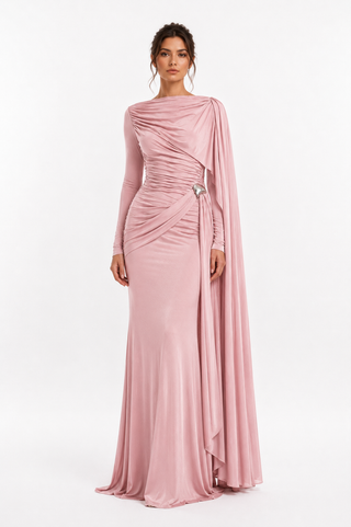 A woman modeling a floor-length dusty pink gown with long sleeves, draped asymmetric detailing, and a flowing cape-like overlay, photographed against a clean white studio background.