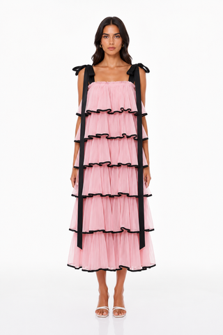 A model wearing a pink tiered tulle dress with black ribbon straps and black ruffle trim, standing against a plain white studio background.