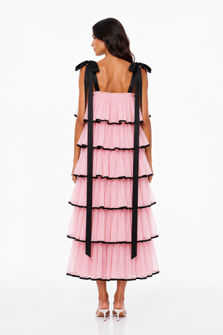 A model viewed from the back wearing a light pink tiered tulle dress with black ribbon shoulder ties and black ruffle trim, paired with white high-heeled sandals against a white studio background.