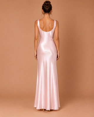 A woman viewed from behind wearing a floor-length satin slip dress in a pale blush pink color, set against a solid warm tan studio backdrop.
