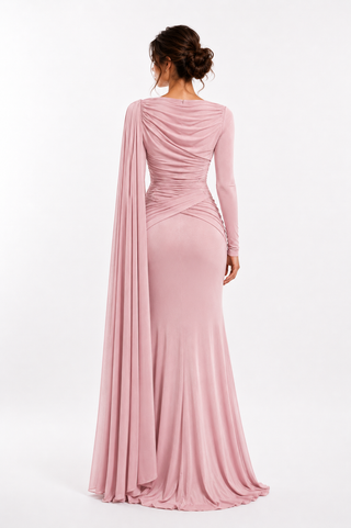A woman shown from the back wearing a floor-length dusty pink gown with long sleeves, draped ruching at the waist, and a flowing cape-like overlay, photographed against a clean white studio background.